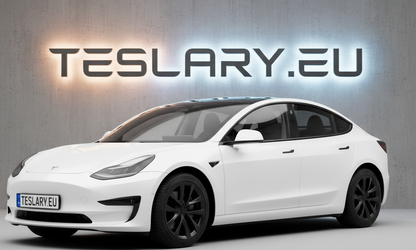 A white Tesla Model 3 sedan shown from a three-quarter front angle, parked in front of a gray concrete wall with a glowing 'TESLARY.EU' sign behind it. tesla-model-3-18-arachnid-style-wheel-covers-teslary-dublin-ireland-cork-nederlands-deutschland-espana-alicante-madrid-tesla-accessories-shop-europe-france-ital-4448983