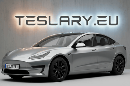 A silver Tesla Model 3 sedan shown from a three-quarter front angle, parked in front of a textured gray wall with a glowing 'TESLARY.EU' sign behind it. tesla-model-3-18-arachnid-style-wheel-covers-teslary-dublin-ireland-cork-nederlands-deutschland-espana-alicante-madrid-tesla-accessories-shop-europe-france-ital-4448983