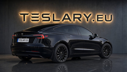 A black Tesla Model Y sedan shown from a rear three-quarter angle in a dark studio setting, with large illuminated TESLARY.EU lettering on the wall behind it. TESLAR MODEL 3 HIGHLAND 18 INCH WHEEL COVERS UBER NOVA PERFORMANCE STYLE GLOSS BLACK FREE DELIVERY WITH FEDEX ACROSS EUROPE FROM IRELAND AND EUROPES TESLARY.EU THE TESLA ACCESSORIES SPECIALISTS